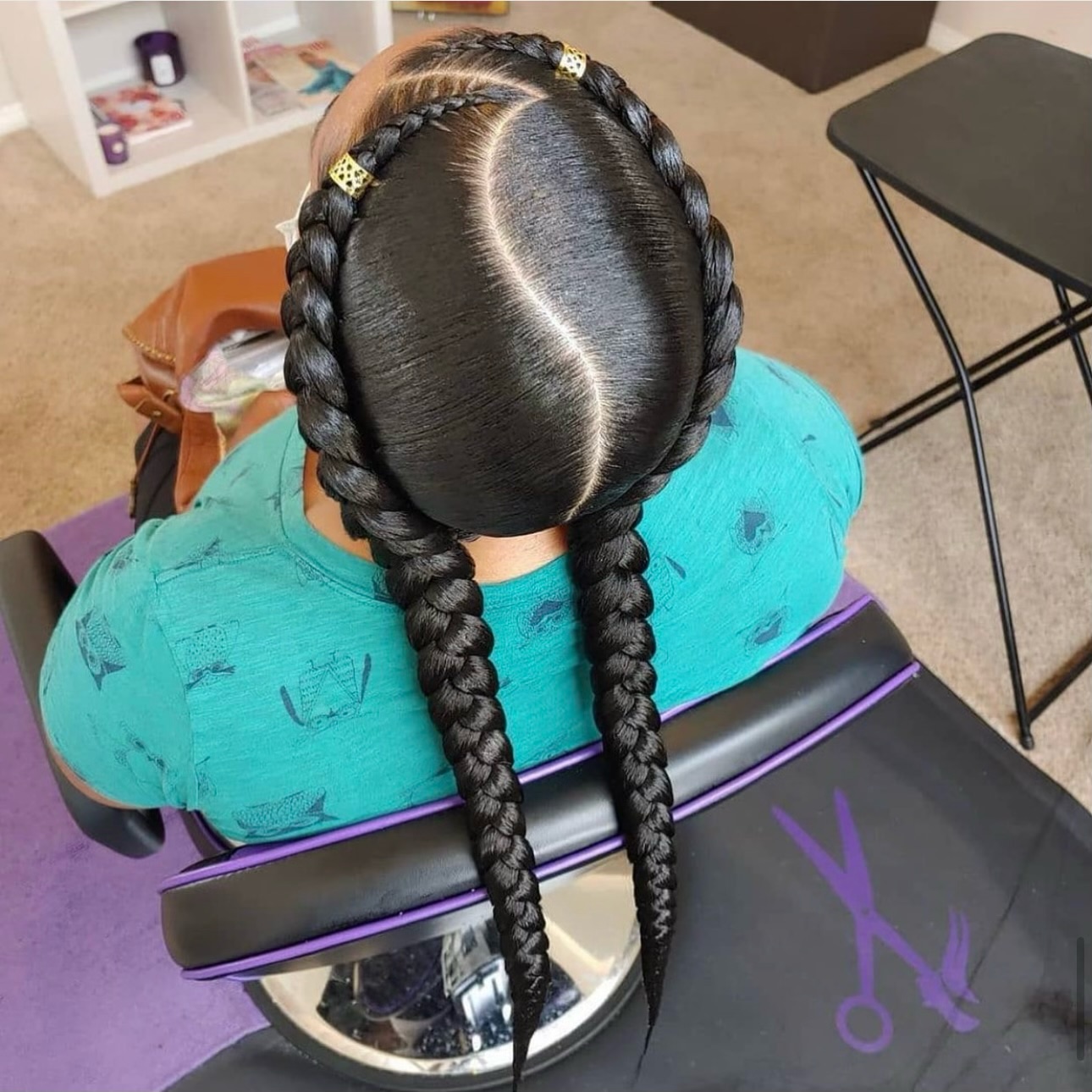 Child with black braided hair styled with gold hair clips, wearing turquoise shirt, seated indoors